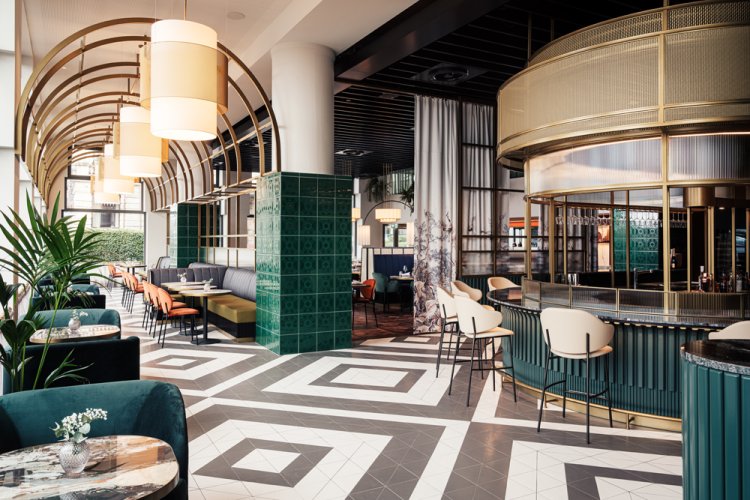 Hotelbar in Dunkelgrün und Gold rechts, im Vordergrund der Boden mit schwarz-weissem Diamantenmuster, links hinten das Restaurant mit Bögen an der Decke, alles im Art déco Stil.