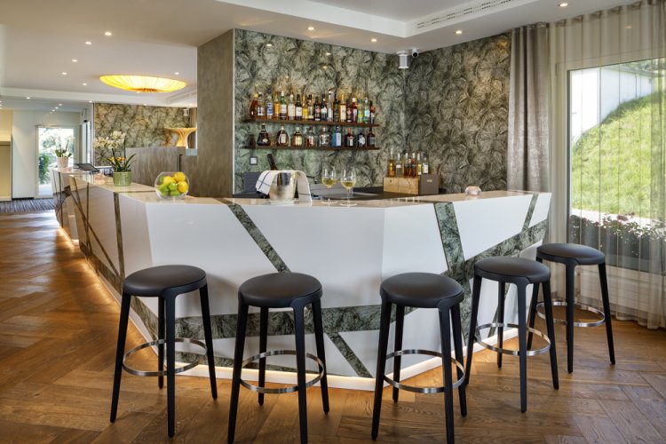 Bar mit Wand aus organischem Argoprint in einem Grünton, durchbrochen mit weissen, grafischen Elementen. Davor schwarze Barhocker auf einem Holzboden.