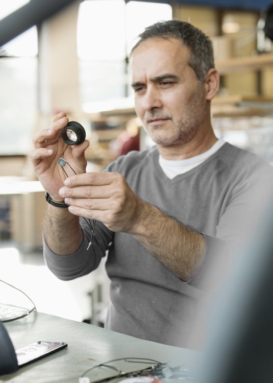 Handwerker in grauem Oberteil schaut Teil einer Leuchte unter der Lupe an