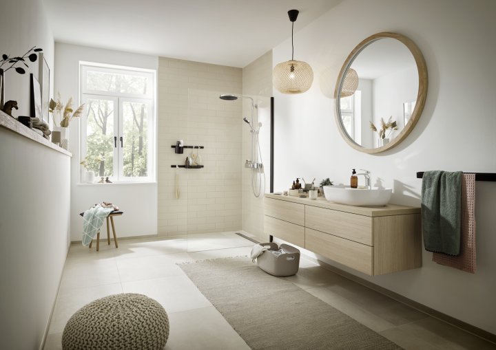 Bad in warmweissen Tönen mit Waschtisch aus hellem Holz mit rundem Spiegel darüber, dahinter eine Dusche mit schwarzen Armaturen und hellen Fliesen.