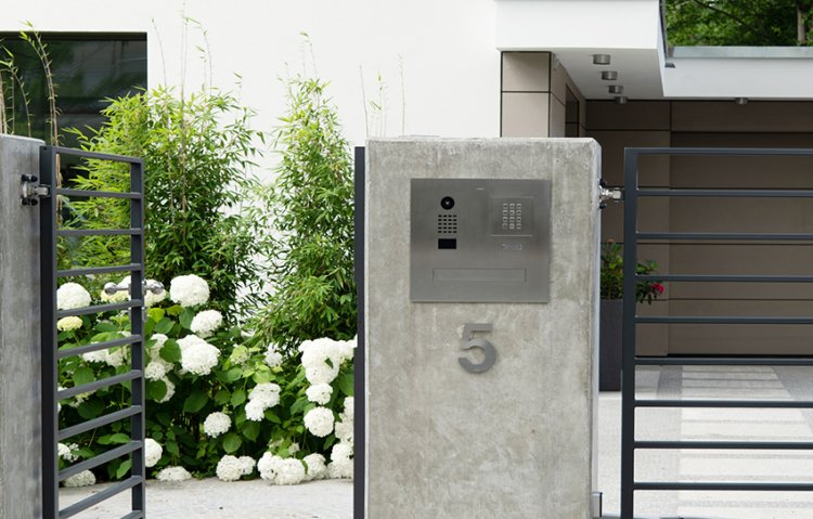Doorbird Schliessystem an Betonsäule mit weissen Blumen und Gartentür im Hintergrund