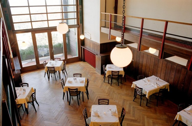 Grosse Halle des Looshauses mit Holzboden und vielen, Quadratischen Esstischen mit weissen Tischtüchern, die als Restaurant funktioniert.