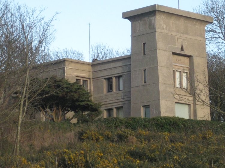 Betongebäude mit einem Turm am vorderen Ende im ägyptischen Stil umgeben von kahlen Bäumen und einigen Büschen im Vordergrund.