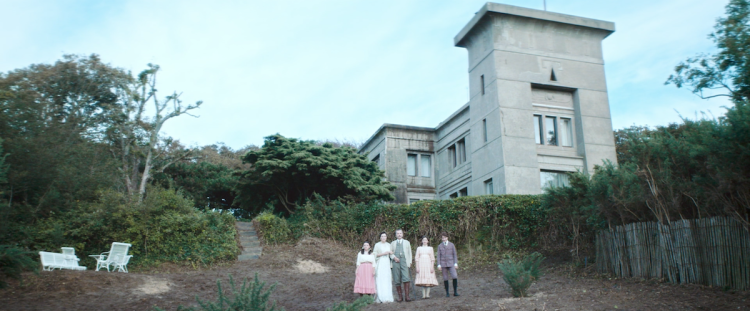 Eine Familie in historischen Kleidern posiert vor einem monumentalen Betongebäude im ägyptischen Stil, umgeben von Büschen und kleinen Bäumen.