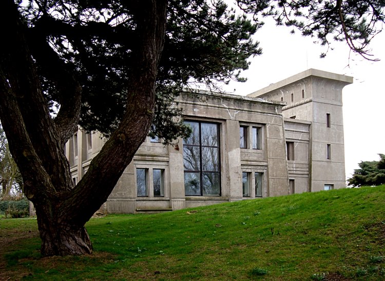 Haus aus Beton angelehnt an den ägyptischen Stil steht momumentartig auf einem Grashügel, links teils von einem Baum im Vordergrund verdeckt.