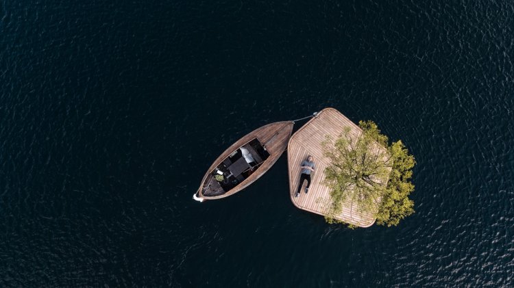 Eine schwimmende Holzinsel mit Baum und angelegtem Boot, auf der ein Mann liegt.