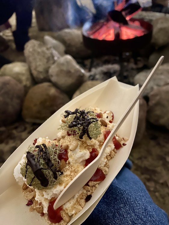 Länglicher Teller mit Dessert auf einem Bein einer Person, die am Lagerfeuer sitzt