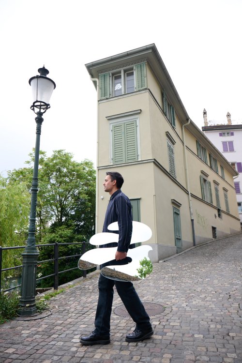 Christian Neuenschwander geht eine gepflasterte Straße in einer Altstadt hinauf. Er trägt eine grosse, organisch geformte Spiegelskulptur unter dem Arm, in der sich die umliegenden Gebäude und der Himmel reflektieren. Im Hintergrund ist ein schmales, helles Gebäude und eine klassische Strassenlaterne zu sehen.