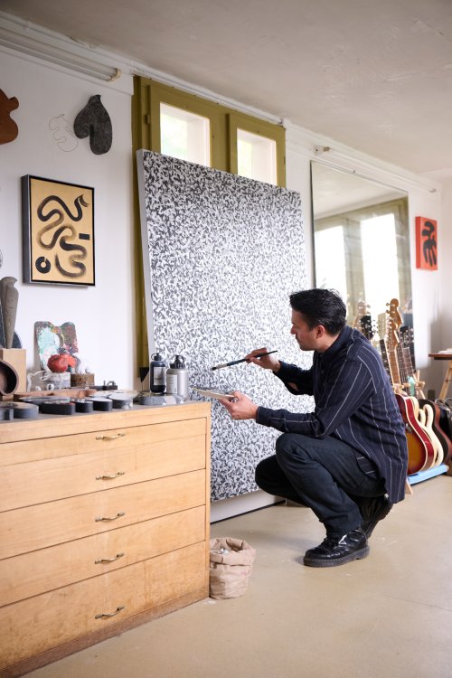 Christian Neuenschwander kniet in seinem Atelier vor einer großformatigen Leinwand und arbeitet mit einem Pinsel an einem detailreichen, schwarz-weissen Werk. Das Bild zeigt dichte, punktierte oder gesprenkelte Strukturen, die organische Formen bilden. Im Hintergrund lehnen mehrere Gitarren an der Wand.
