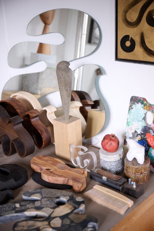 Nahaufnahme eines Arbeitstisches im Atelier von Christian Neuenschwander. Zu sehen sind verschiedene skulpturale Objekte aus Holz und Stein, eine farbbekleckerte Palette, ein Granatapfel auf einem Betonsockel und im Hintergrund ein wellenförmig geschnittener Spiegel, der den Raum reflektiert.