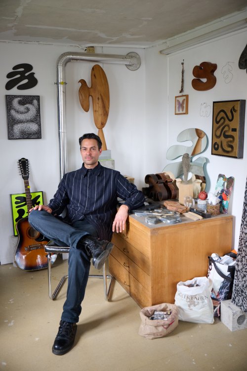 Ein Porträt von Christian Neuenschwander, der entspannt auf einem Stuhl in seinem hellen Atelier sitzt. Um ihn herum befinden sich seine Werke, darunter Holzskulpturen, Gemälde mit abstrakten Formen und eine Gitarre. Der Raum wirkt wie eine kreative Werkstatt mit einer Mischung aus Kunstobjekten und Arbeitsmaterialien.