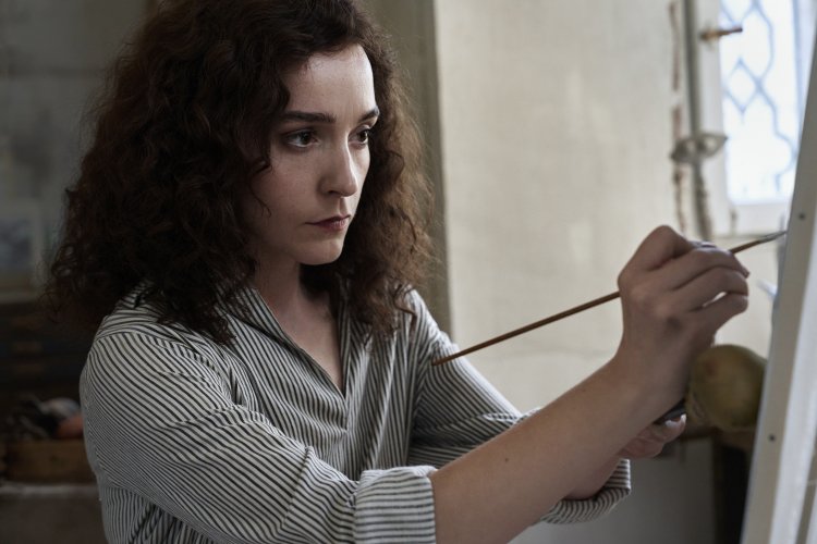 Filmstill aus Leonora in the Morning Light: Eine Frau mit lockigem Haar malt konzentriert mit einem feinen Pinsel an einer Staffelei in warmem Licht.