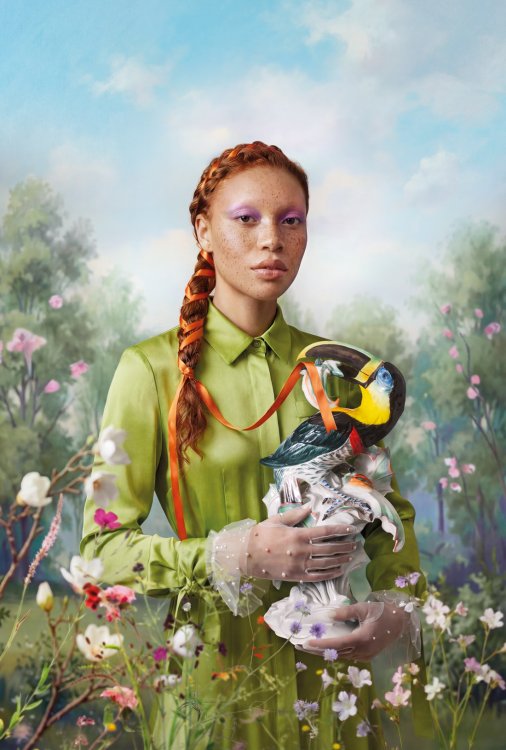 Porträt einer Frau im grünen Kleid, die eine farbige Papageienfigur inmitten einer blühenden Frühlingslandschaft hält.