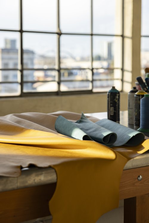 Lederbahnen in verschiedenen Farben liegen in einem Atelier auf einem Arbeitstisch vor grossen Fenstern; daneben stehen Farbdosen und Garnrollen.
