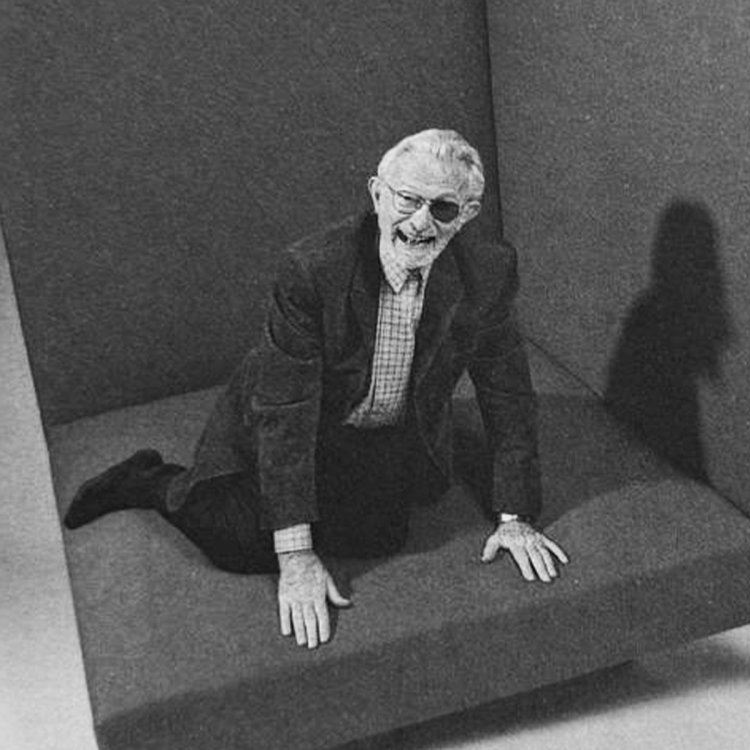 Schwarz-Weiss-Porträt des Schweizer Designers Dieter Waeckerlin, der lächelnd auf einem grossen, gepolsterten Sitzelement kniet.