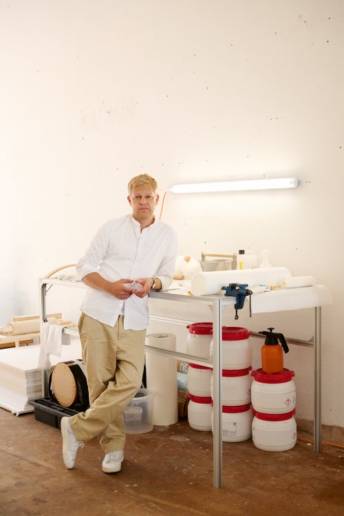 Ein Porträt des Designers Ville Kokkonen in seinem hellen Atelier. Er steht entspannt mit verschränkten Beinen an einem Arbeitstisch aus Metall und trägt ein weisses Hemd sowie beige Hosen. Der Hintergrund zeigt eine minimalistische Werkstattumgebung mit weissen Wänden, einer Leuchtstoffröhre und verschiedenen Arbeitsmaterialien wie Papierrollen, weissen Behältern mit roten Deckeln und Werkzeugen.