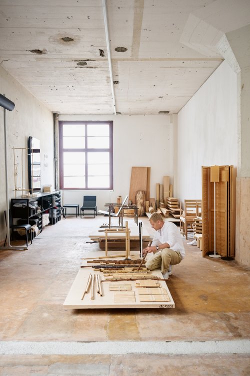 Der Designer Ville Kokkonen kniet in seinem grossräumigen, lichtdurchfluteten Atelier auf dem Boden und begutachtet verschiedene Holz- und Bambuselemente, die auf einer grossen Platte ausgelegt sind. Der Raum im industriellen Stil zeigt im Hintergrund weitere Prototypen, Holzstapel und ein grosses Fenster, was die kreative Arbeitsatmosphäre unterstreicht.