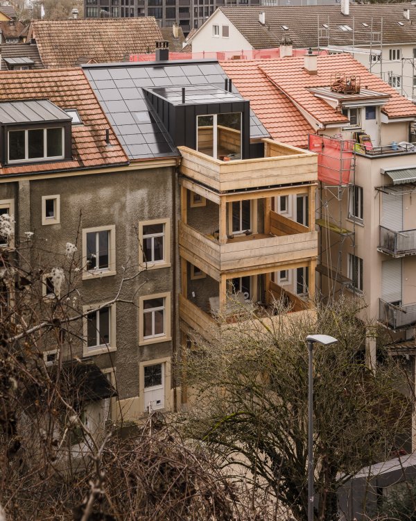 Aussenansicht eines renovierten, mehrgeschossigen Arbeiterhauses in Münchenstein. Zu sehen ist ein neuer, dreigeschossiger Balkonanbau aus hellem Massivholz und ein modernisiertes Dach mit dunkler Photovoltaik-Eindeckung und einer schwarzen Dachlukarne. Das Gebäude steht in einer dichten städtischen Nachbarschaft.