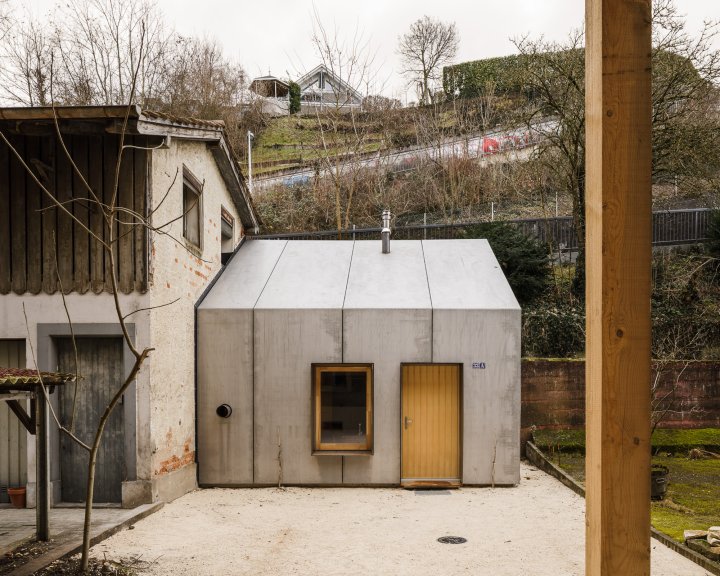Frontansicht eines minimalistischen Tiny Houses im Hinterhof eines Altbaus in Münchenstein. Das kleine Gebäude mit Satteldach ist vollständig mit hellgrauen Stahlpaneelen verkleidet. In der Fassade befinden sich ein holzgerahmtes Fenster mit metallener Laibung und eine schlichte Holztür. Links grenzt direkt die verputzte Fassade eines älteren Werkstattgebäudes an.