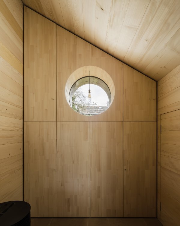 Innenansicht des Tiny Houses mit raumhohem Ausbau aus hellem Nadelholz. Die Wände und die schräge Decke sind mit Holzpaneelen verkleidet, die eine warme Atmosphäre schaffen. In der Stirnwand ist ein kreisrundes Fenster eingelassen, davor hängt eine schlichte Glühbirne an einem schwarzen Kabel. Das Holzbild zeigt eine präzise vertikale Linienführung der Einbauschränke.