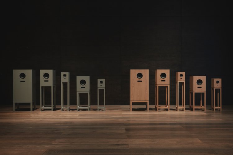 Zehn verschiedene Lautsprecher-Modelle von OJAS und Karimoku in unterschiedlichen Grössen und Holzausführungen (Pewter Grey und Eiche Natur) sind in einer Reihe auf dem dunklen Holzboden des Karimoku Research Centers aufgereiht. Die symmetrische Anordnung vor einer dunklen Wand erinnert an einen physischen Produktkatalog.