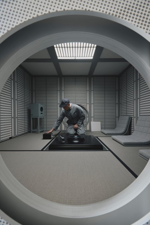 Blick durch eine kreisförmige Wandöffnung in ein modern interpretiertes japanisches Teehaus mit Tatami-Matten, Akustikpaneelen und grauen Bodenstühlen. Im Zentrum kniet Audiodesigner Devon Turnbull an einem schwarzen Plattenspieler-Modul und bedient die Technik.