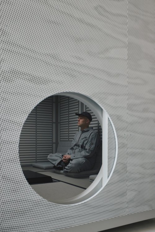 Seitenansicht durch eine kreisförmige Wandöffnung: Audiodesigner Devon Turnbull sitzt im Profil meditativ im Schneidersitz auf einem grauen Bodenstuhl im «Sound House». Im Hintergrund sind horizontale Holzlamellen und eine reduzierte, grau-beige Ästhetik zu sehen.