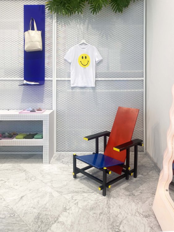 Ein moderner, geometrischer Holzstuhl in Rot, Blau und Gelb steht auf einem hellen Marmorboden. Dahinter befindet sich eine halbtransparente weisse Gitterwand, durch die bunte Stapel von Kartons schimmern. An der Wand hängt ein weisses T-Shirt mit einem gelben Smiley.