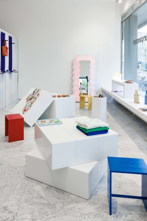 Innenansicht des «La Garçonne» Stores mit Fokus auf die Möblierung: Im Vordergrund stehen bunte Max Bill Hocker und weisse, kubische Präsentationsmodule auf Marmorboden. Im Hintergrund lehnt der ikonische, rosafarben leuchtende «Ultrafragola» Spiegel von Ettore Sottsass an der weissen Wand.