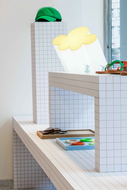 Eine weiss leuchtende Lampe in Form einer stilisierten Wolke mit gelber Kontur steht auf einem weissen Tisch mit schwarzem Karomuster. Auf dem Tisch liegen zudem bunte Armreifen und Sandalen. Im Hintergrund ist eine grüne Baseballkappe auf einer hohen, ebenfalls karierten Säule zu sehen.