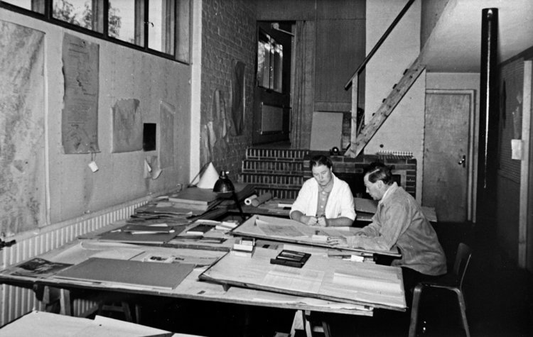 Historische Schwarz-Weiss-Fotografie von Aino und Alvar Aalto bei der Arbeit in ihrem Architekturbüro. Sie sitzen nebeneinander an grossen Zeichentischen, die mit Entwürfen und Dokumenten bedeckt sind, in einem hellen Atelier mit hohen Fenstern und Backsteinwänden.