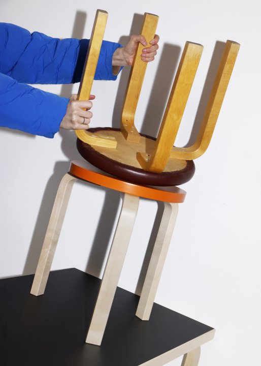 Zwei aufeinandergestapelte Artek-Hocker von Alvar Aalto. Eine Person in einer blauen Daunenjacke hält den oberen Hocker, der kopfüber auf einem orangefarbenen Hocker steht, welcher wiederum auf einem schwarzen Tisch platziert ist.
