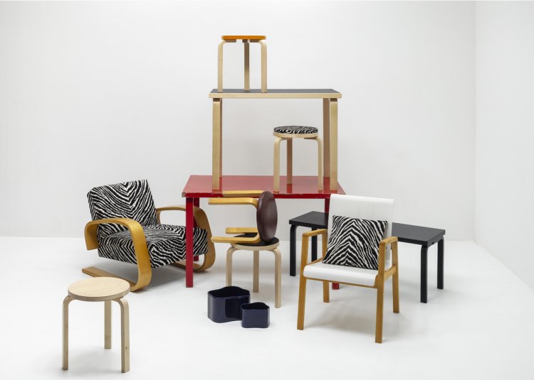 Verschiedene Artek-Möbelklassiker von Alvar und Aino Aalto sind in einem weissen Raum kunstvoll arrangiert und teilweise gestapelt. Zu sehen sind Sessel mit Zebra-Muster, diverse Hocker 60, ein roter Tisch, eine schwarze Bank sowie dunkelblaue Aalto-Vasen. Das Arrangement wirkt wie eine spielerische Installation in einem Möbellager.