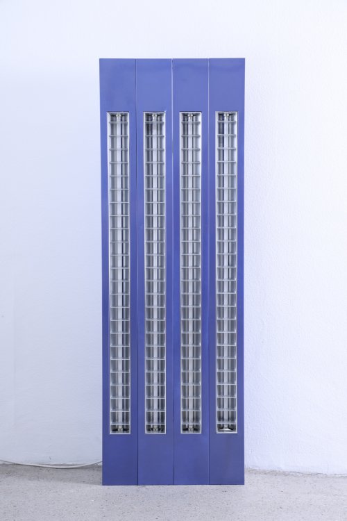 Ein vertikales Designobjekt von Altherr Weiss, bestehend aus vier schmalen, violett-blauen Säulen, in die industriell wirkende Rasterleuchten mit Leuchtstoffröhren integriert sind. Die minimalistische Lichtinstallation steht vor einer neutralen weissen Wand auf einem grauen Betonboden.