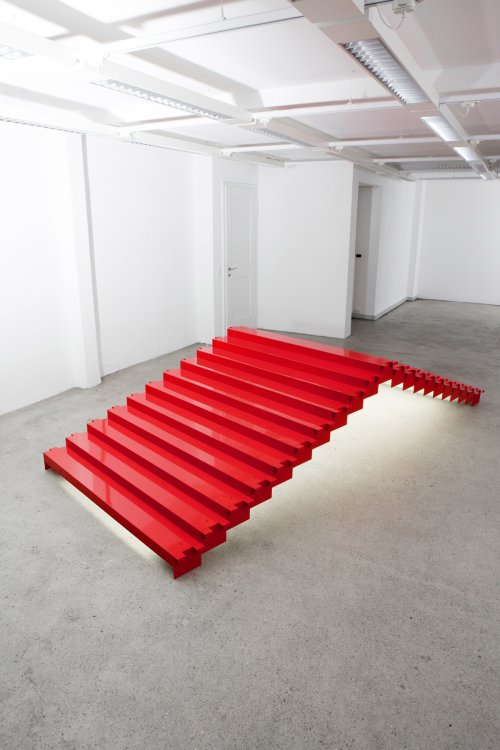Eine auffällige, leuchtend rote Installationsskulptur mit dem Titel „Pyramid“ von Altherr Weiss in einem minimalistischen Gallerieraum. Das Objekt gleicht einer breiten, flach ansteigenden Treppe aus Metallsegmenten, die im hinteren Bereich steil abfällt und von unten indirekt beleuchtet wird, sodass sie über dem Betonboden zu schweben scheint.