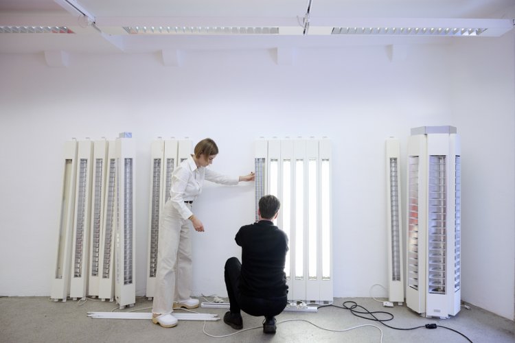 Das Designduo Altherr Weiss bei der Arbeit in einem hellen, weissen Raum, umgeben von mehreren vertikalen, industriell anmutenden Leuchtelementen. Eine Frau in heller Kleidung steht links und justiert eine der Leuchten an der Wand, während ein Mann in schwarzer Kleidung davor kniet und eines der hell leuchtenden Objekte betrachtet.