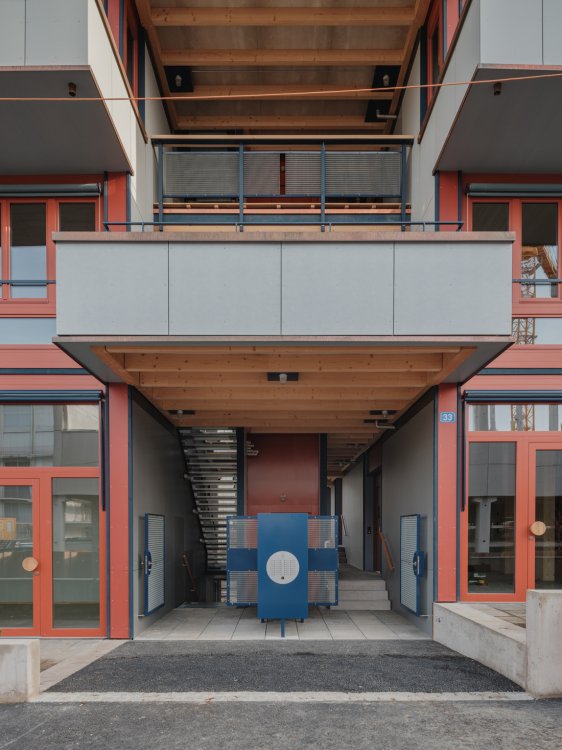 Blick in den überdachten Eingangsbereich des Wohnhauses LysP8. Sichtbare Holzbalken an der Decke und rote Fensterrahmen prägen die Ansicht; im Zentrum steht ein blaus Element mit Briefkästen vor einer Treppe, darüber ragen Balkone mit grauen Brüstungen hervor.