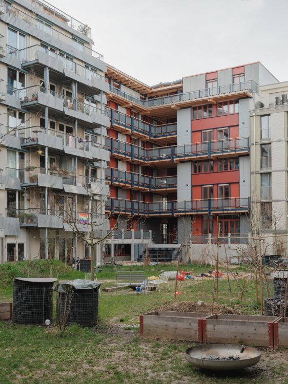 Blick aus dem begrünten Innenhof auf die rückwärtige Fassade des Wohnhauses LysP8. Charakteristisch sind die umlaufenden Laubengänge aus hellem Holz mit dunklen Metallgeländern sowie fassadenhohe rote Wandelemente. Im Vordergrund sind Hochbeete, eine Feuerschale und ein naturnah gestalteter Gartenbereich zu sehen.