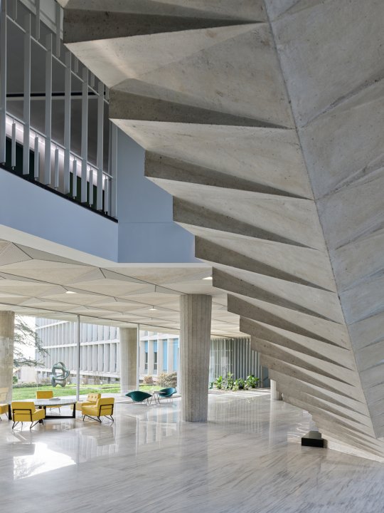 Skulpturale Wendeltreppe aus Sichtbeton in der lichtdurchfluteten Eingangshalle des «Le Cèdre» in Lausanne. Die geometrische Untersicht der Treppe bildet einen markanten Kontrast zum hellen Marmorboden, den filigranen Betonsäulen und den gelben Designer-Sesseln im Hintergrund.