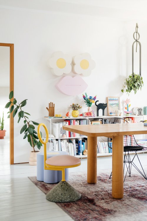 Heller Wohnraum im Pop-Art-Stil mit einem grossen Wandobjekt in Form eines Gesichts, bestehend aus zwei Gänseblümchen als Augen und einem rosa Kussmund. Im Raum stehen ein massiver Holztisch, ein verspielter gelb-rosa Stuhl sowie Design-Hocker.