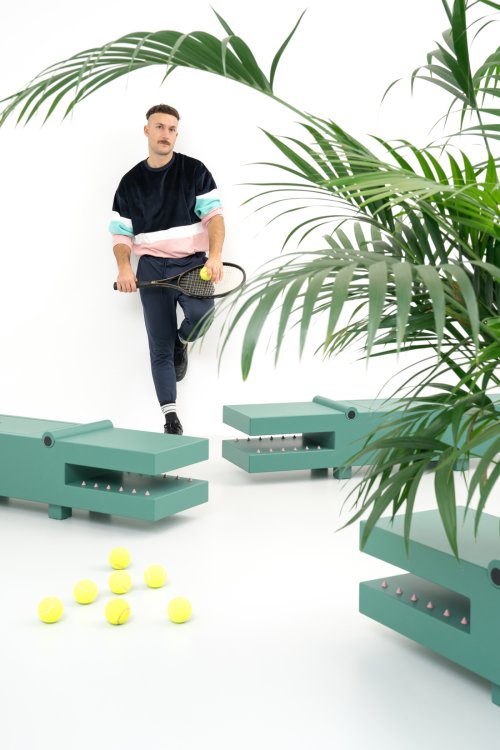 Ein modisch gekleideter Mann mit Schnurrbart lehnt mit einem Tennisschläger an einer weissen Wand, umgeben von surrealen, grünen Sitzmöbeln in Form von Krokodilen und verstreuten gelben Tennisbällen. Im Vordergrund sind grüne Blätter einer Palme zu sehen, die dem minimalistischen Studio-Setting eine frische Atmosphäre verleihen.
