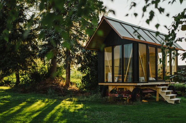 Mobiler Pavillon aus dunklem Holz mit steilem Satteldach und grossflächiger Verglasung auf einer Wiese im Abendlicht. Die Holzkonstruktion ruht auf einem Fahrgestell mit Rädern, eine kleine Holztreppe führt zum Eingang.