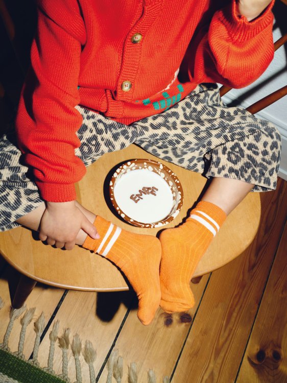 Draufsicht auf ein Kind, das auf einem Holzstuhl sitzt. Es trägt einen leuchtend roten Strickpulli, Hosen mit Leopardenmuster und orangefarbene Socken. In der Mitte steht eine braun-weiss gesprenkelte Emaille-Schale (Mirari Bowl) von ÉMAÉ STUDIO, in deren Milchfüllung Schokobuchstaben das Wort „EMAE“ bilden.