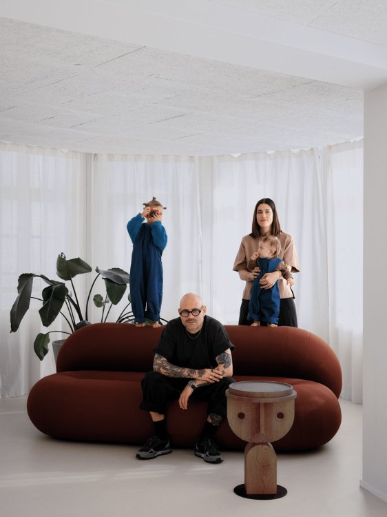 Porträt der Gründerfamilie von ÉMAÉ STUDIO: Nadia Studer mit Baby auf dem Arm, ihr Mann Ramon Studer auf einem rötlichen Design-Sofa sitzend und ihr älterer Sohn, der hinter dem Sofa mit einer Kamera spielt. Helles Interieur mit grossen Fenstern und weissen Vorhängen.