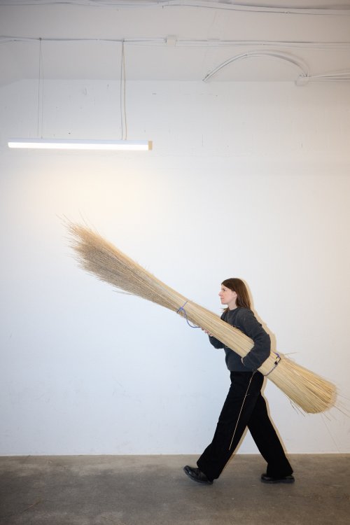 Designerin Flavia Brändle, gekleidet in Schwarz, trägt ein sehr grosses Bündel aus langen Naturfasern durch einen hellen Raum.