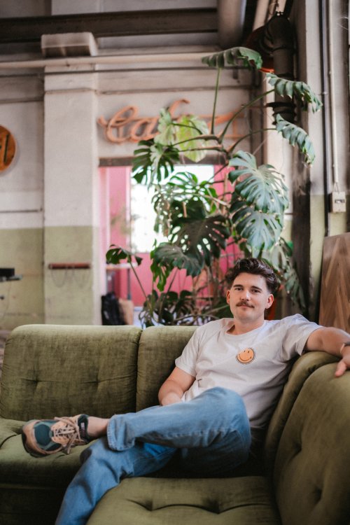 Ein Porträt von Marius Gierhardt, dem Gründer von Fisch&Tisch, der entspannt auf einem olivgrünen Samtsofa in einer Industriehalle sitzt. Im Hintergrund eine grosse Monstera-Pflanze und ein rustikaler «Café»-Schriftzug.