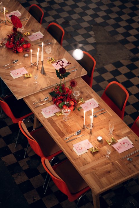 ine lange, festlich gedeckte Tafel aus Fischgrätparkett-Tischen in einer Industriehalle mit schwarz-weiss kariertem Boden. Die Tische sind mit brennenden Kerzen in silbernen Leuchtern, rotem Blumenschmuck, Weingläsern und roten Designerstühlen arrangiert.