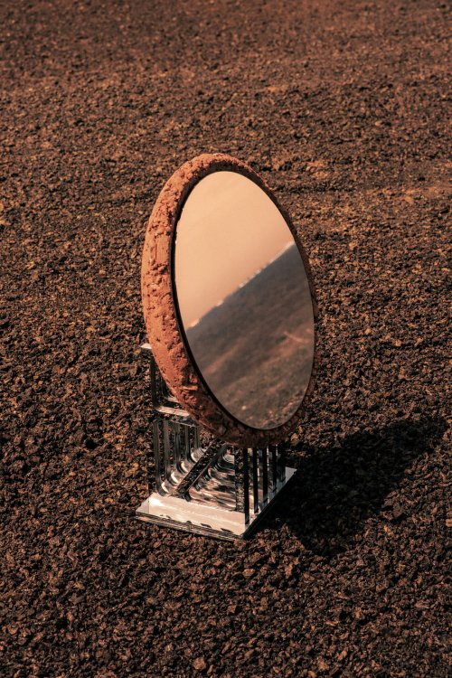 Faded Signal: Ovale, spiegelnde Scheibe mit rauem Stampflehmrand auf einer filigranen Edelstahlbasis, auf einer dunklen, vulkanisch wirkenden Oberfläche fotografiert.