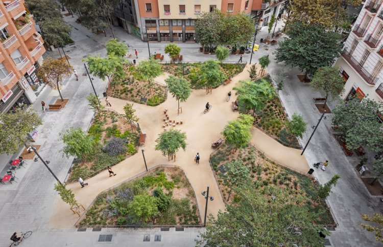 Luftaufnahme eines umgestalteten Strassenkreuzes in Barcelona: Ein grosser, sternförmiger Platz mit hellen Kieswegen und dichten, grünen Beeten ersetzt den Asphalt, umgeben von Wohnhäusern.