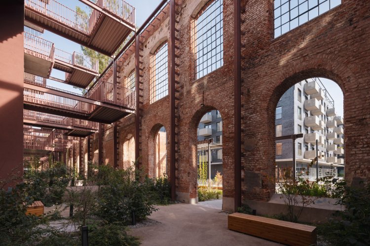 Innenhof eines revitalisierten Industrieareals mit historischen Backsteinmauern und grossen Bogenfenstern, daneben moderne Stahlgänge und Holzbänke.